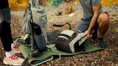 Close-up-shot-of-a-girl-and-a-guy-disassembling-their-backpacks-and-taking-out-equipment-for-rock-climbing-on-a-rocky-beach-near-the-yellow-rocks-during-the-day-near-the-sea