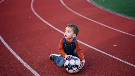 little boy follows the ball rolling by the ground. cute child plays at the stadium. teenage football players at backdrop.