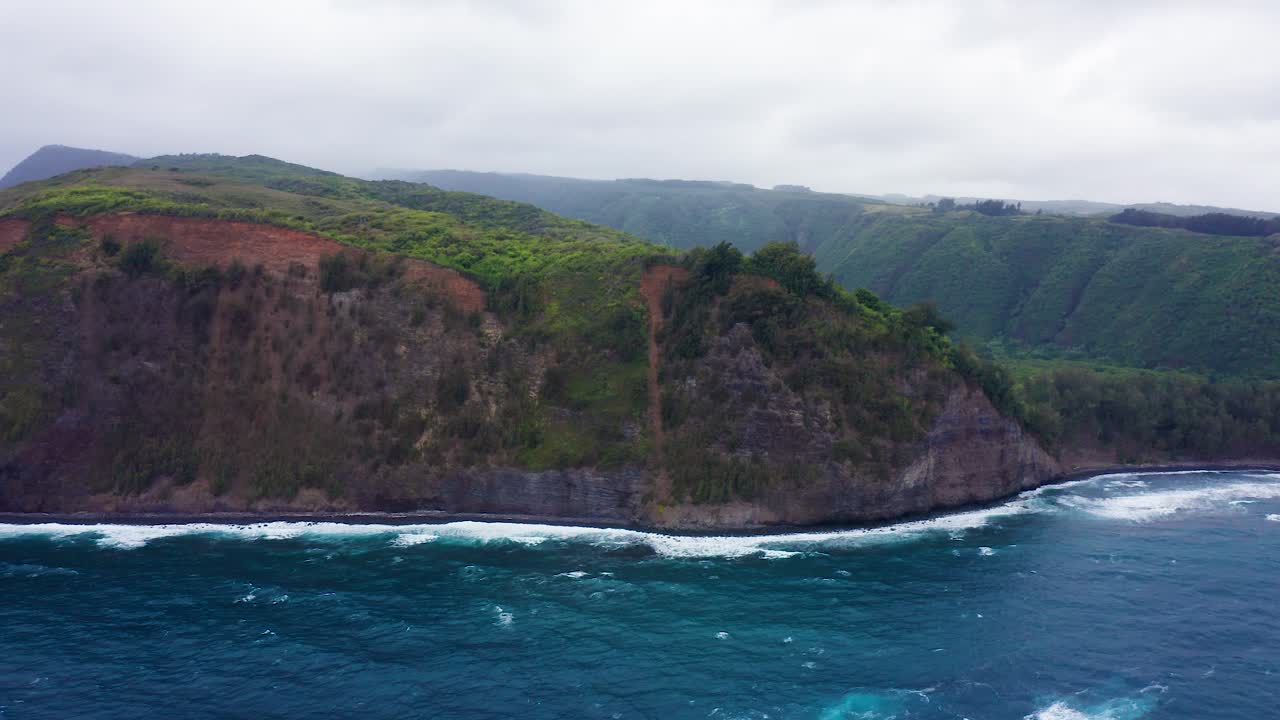 In Polulu Valley, Hawaii, dramatic cliffs capped with green and red earth plunge to a restless deep blue ocean where waves break along the volcanic shore beneath overcast skies.