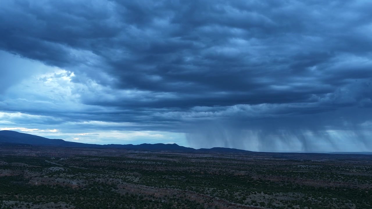 4K drone-timelapse som fanger mørke regnstriper som feier over et vidstrakt ørkenplatålandskap under blåtime. Tunge, stemningsfulle solnedgangsskyer truer over det ulendte terrenget i et kjølig kveldslys.