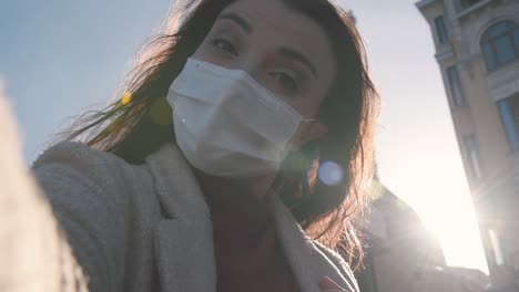 beautiful girl wearing protective medical mask and fashionable clothes takes selfie with a smart phone at street