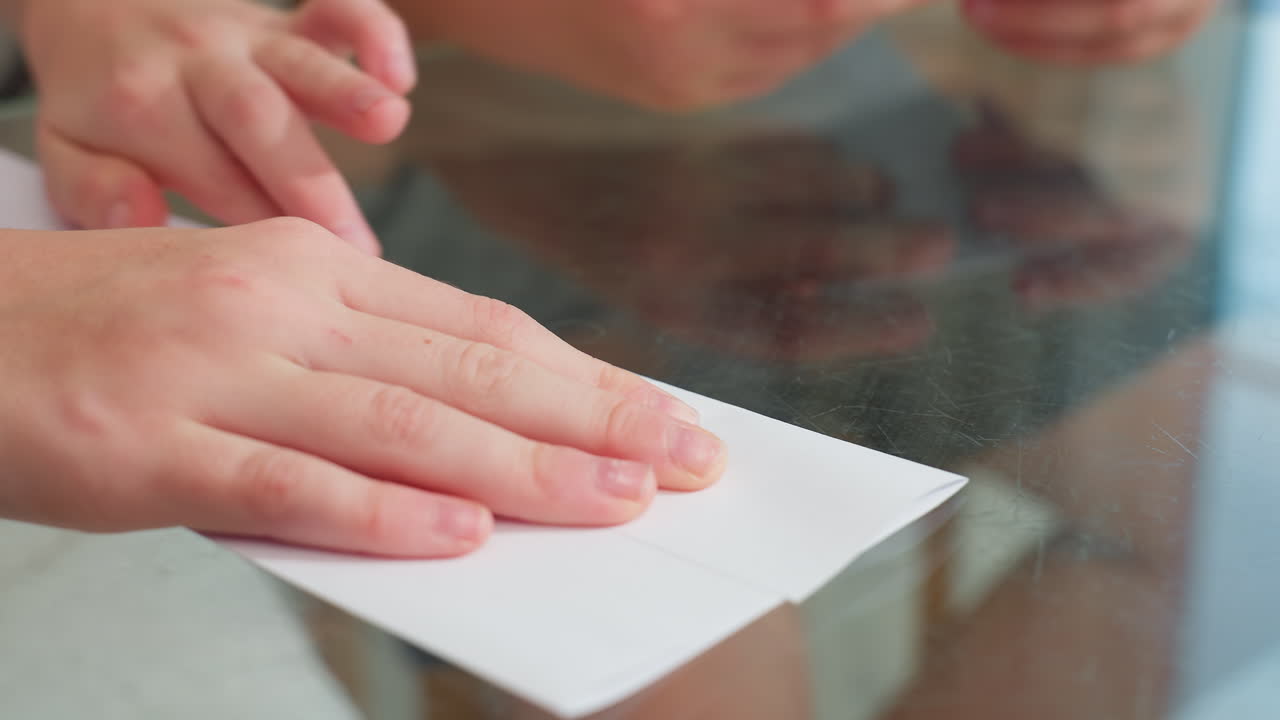 Close-up of hands bending and straightening paper craft on glass table, carefully aligning edges for neatness and precision in crafting process