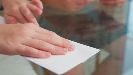 close-up of hands bending and straightening paper craft on glass table, carefully aligning edges for neatness and precision in crafting process