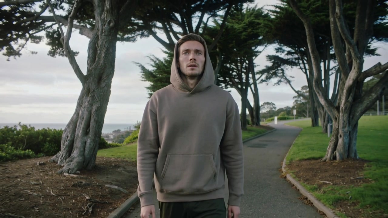 Seorang pria ber-hoodie berjalan mendekati kanopi sambil melambat, lalu berhenti di jalur pantai mengamati ke atas.