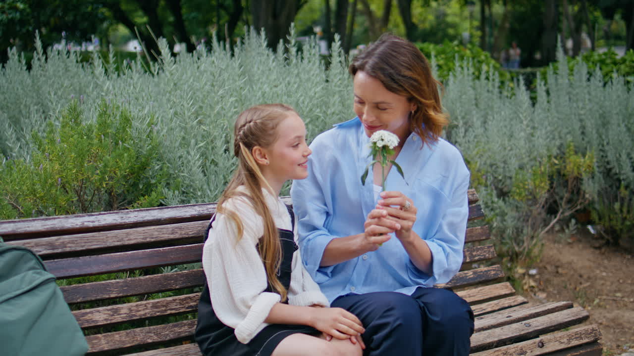 Cute daughter bringing flower to mommy greenery park back view. Family embracing