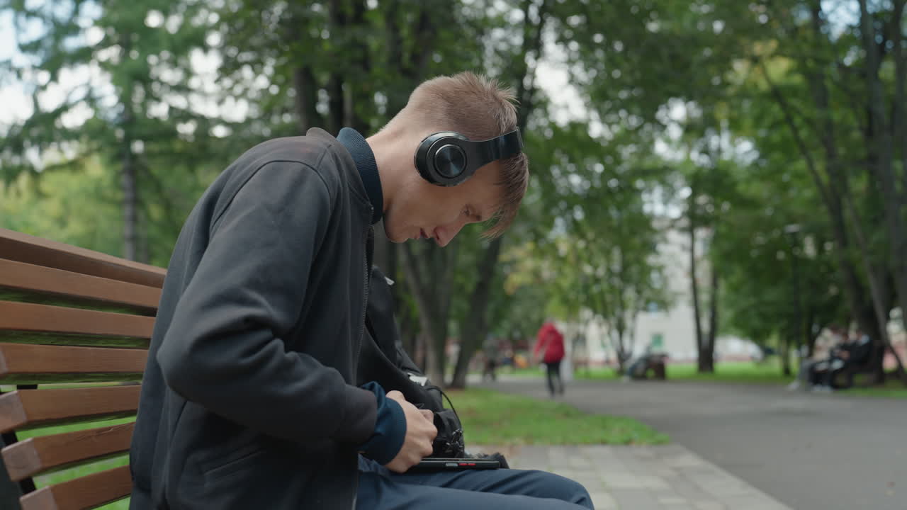 Ung man på bänk som fixar med hörlurar, justerar kabel och portabel ljudenhet medan han bär over-ear-hörlurar, parkträd och asfalterad gångväg i bakgrunden, fokuserad hållning och försiktiga händer