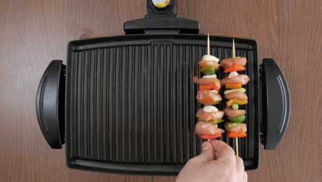 man putting wooden skewers with raw chicken and vegetables on a compact electric grill