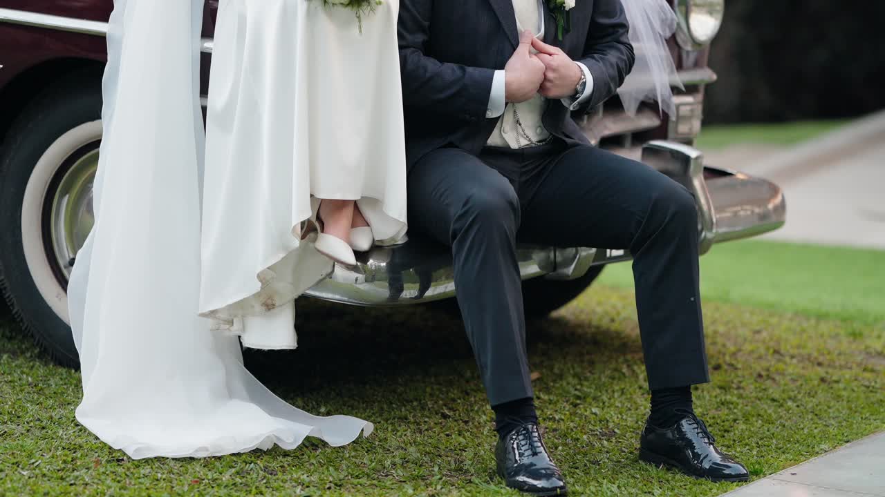 wedding couple sitting together on vintage car bumper with tender hand hold