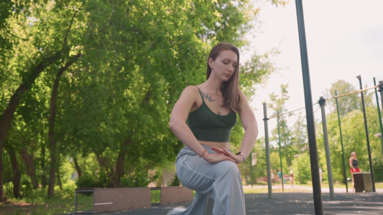 Primo piano di una donna che esegue esercizi di stretching all'aperto alla luce del giorno, con alberi e attrezzature del parco visibili. La donna indossa abbigliamento sportivo che include canotta, pantaloni della tuta, scarpe da ginnastica e orologio.