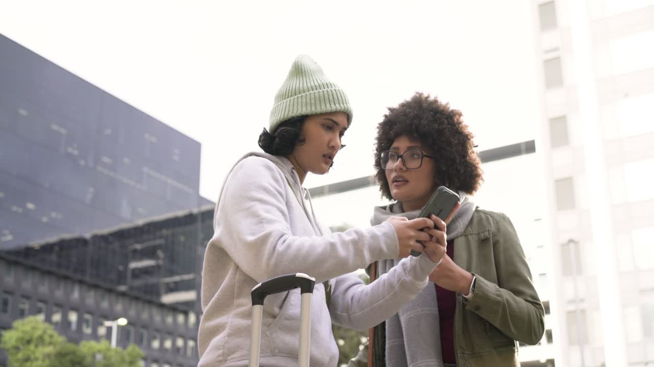 Diverse female friends opening smartphone map on sidewalk with suitcase pointing route and walking