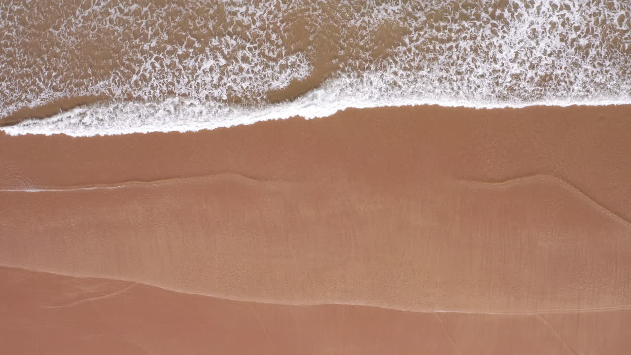 Statisk luftfoto af havbølger, der slår mod en sandet gylden strand.