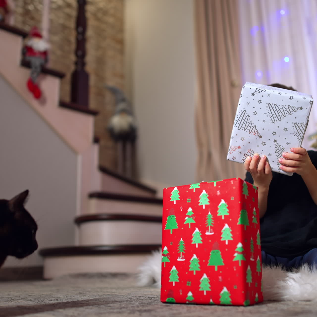 Little kid sitting on the floor near the Christmas tree. Adorable baby boy playing with gift boxes. Black cat watches the child.