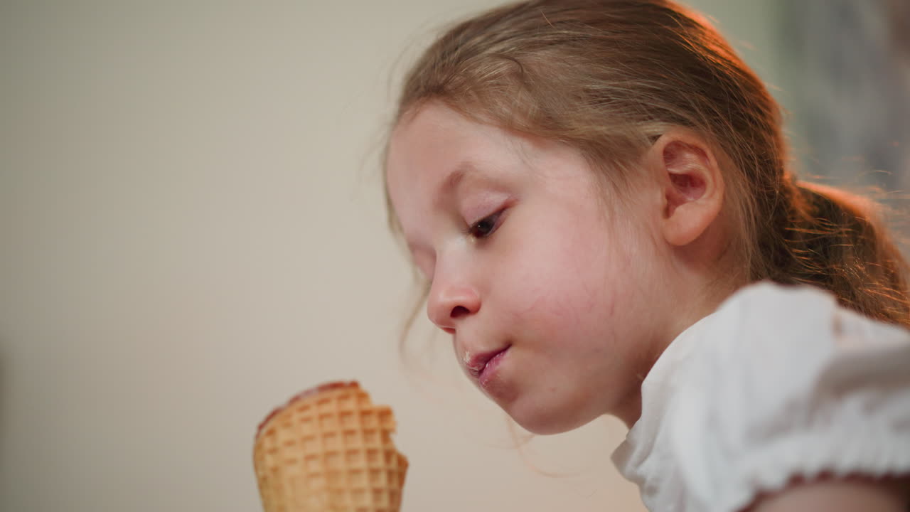 Upward view of young girl enjoying ice cream cone, licking sweet treat with soft smile, casual indoor setting, sunlight highlighting hair, creating warm joyful moment of childhood