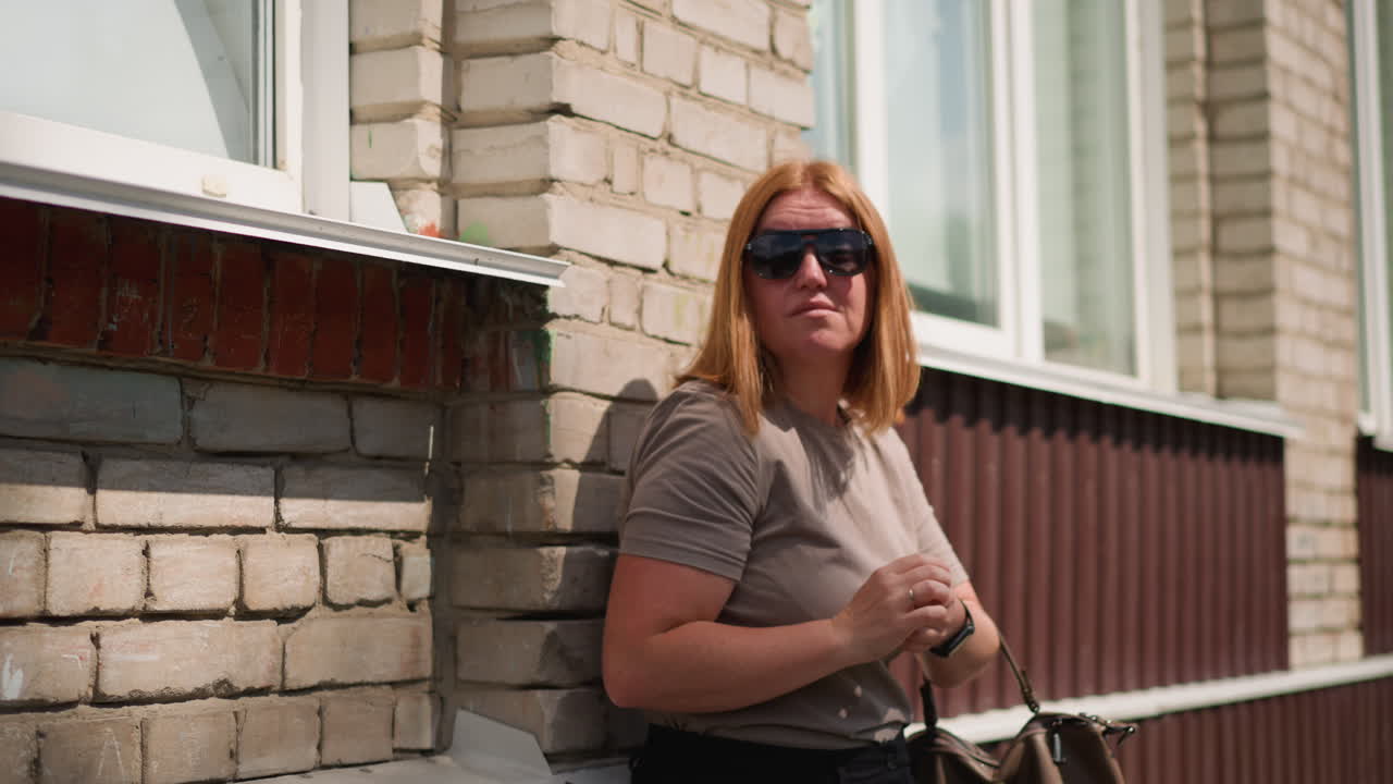 Studerende med lysebrunt hår læner sig op ad en murstensvæg på en solrig eftermiddag, holder en håndtaske og solbriller og kigger til højre og venstre, sollys reflekteres på huden