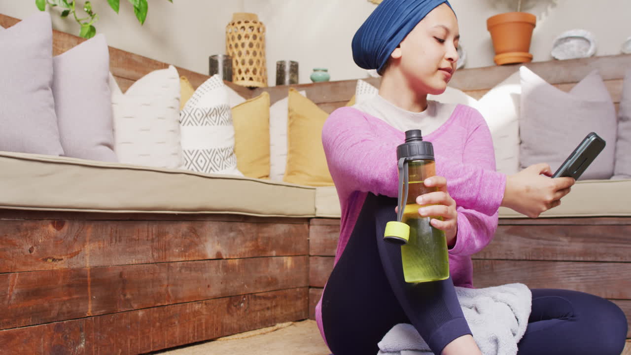 휴게실에서 스마트폰을 사용하여 요가 매트에 앉아있는 히자브를 입은 미소 짓는 두 인종 여성의 비디오