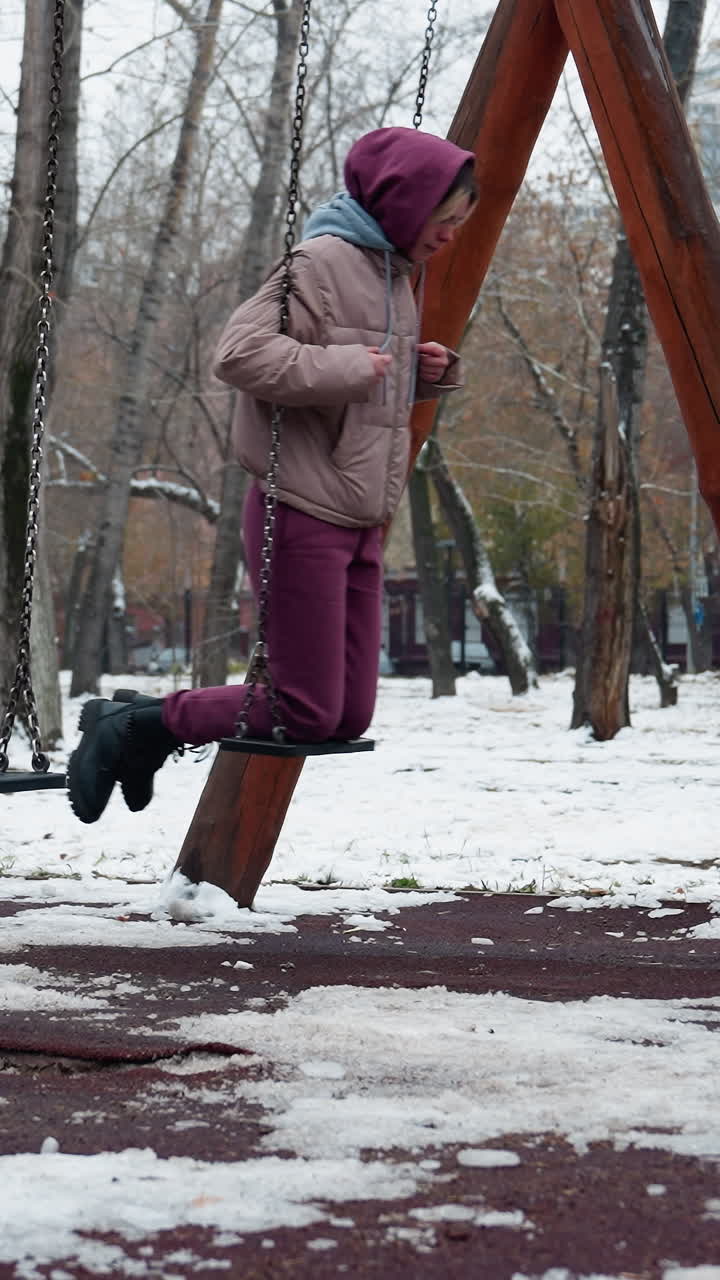 vista lateral de una dama vestida para el invierno con pantalones de color marrón en un columpio sosteniendo cadenas con nieve en el suelo y árboles desnudos en el fondo, disfrutando de actividades al aire libre en clima frío