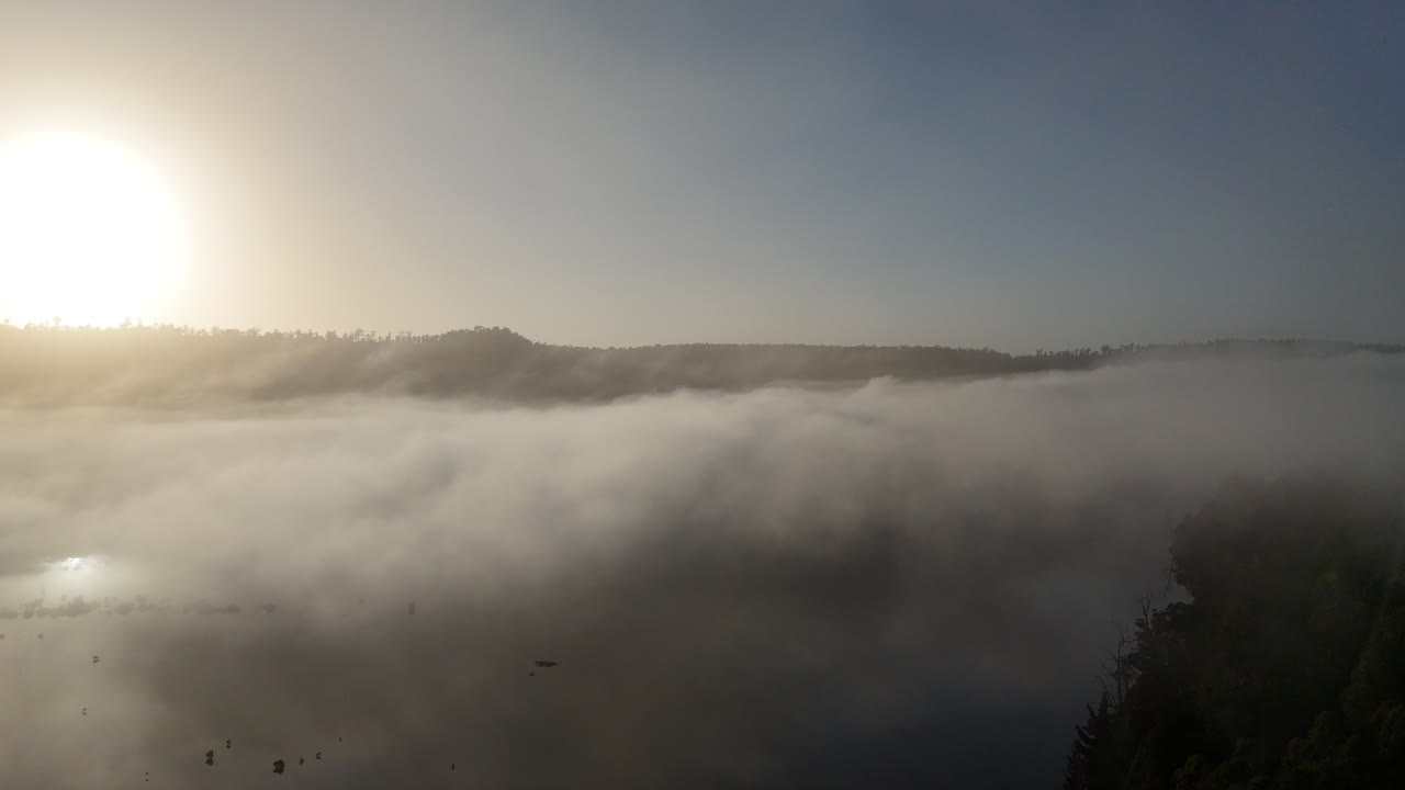 Witness a breathtaking sunrise breaking through clouds as a drone ascends over a tranquil landscape, revealing the beauty of dawn in the mist.