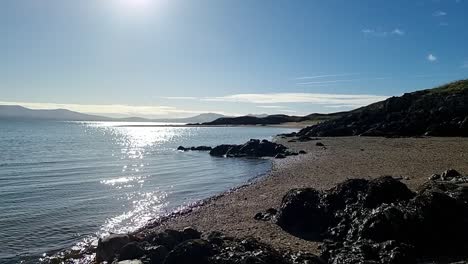 Slow-pan-across-rocky-Anglesey-coastline-with-sunrise-shimmering-ocean-water-and-Snowdonia-mountain-range-horizon