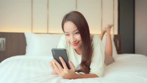 a happy young woman lying on her stomach on the bed taps a message on her cell phone