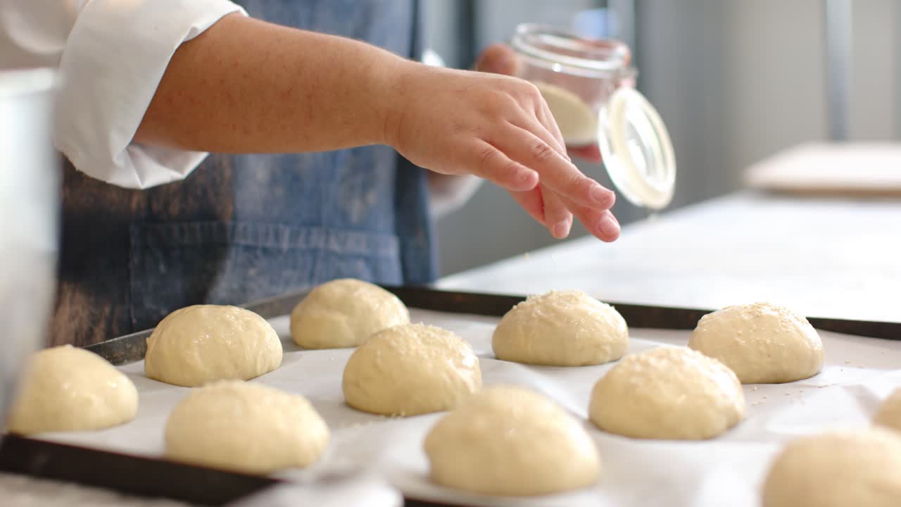Woman in apron placing jar on trays sprinkling seeds over buns for baking at kitchen counter