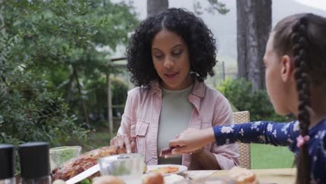 Happy-biracial-mother-and-daughter-eating-meal-at-dinner-table-in-garden,-slow-motion