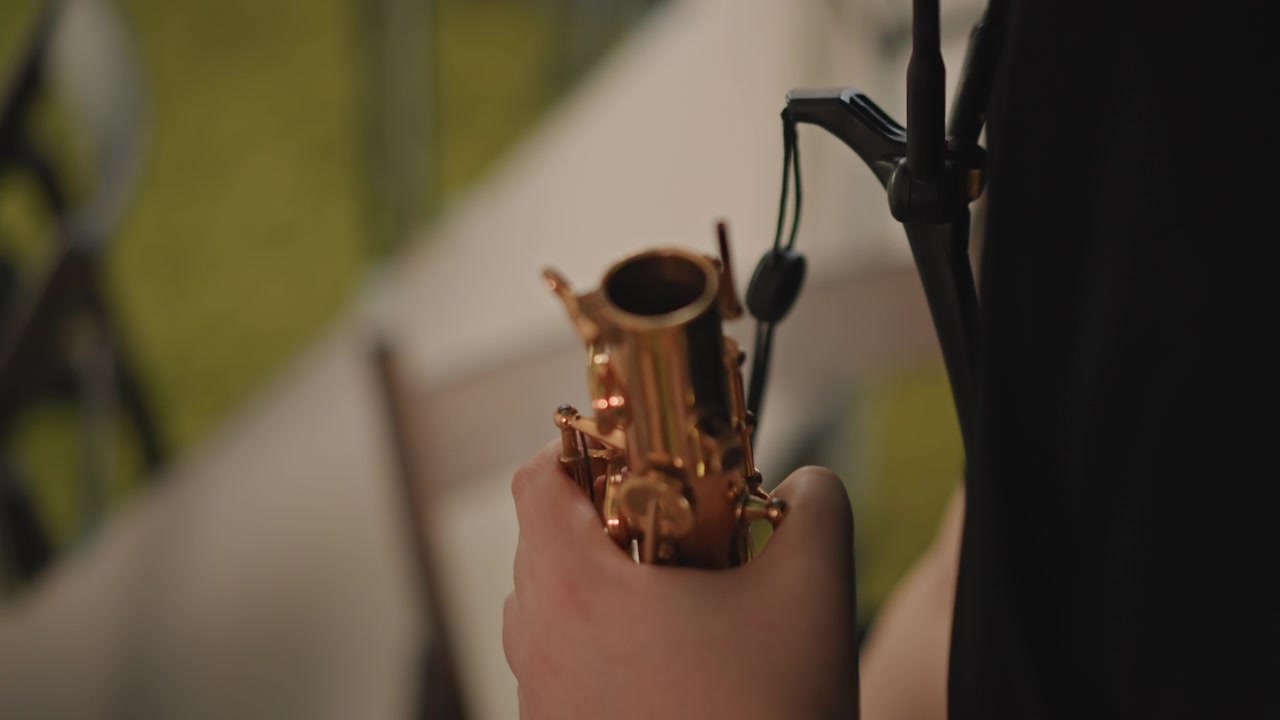 屋外でサックスのマウスピースを外す白人学生のクローズアップ。折りたたみ椅子に座りリハーサルに備える彼の、暖かく金色に輝く真鍮が日光を反射し、手は安定している。近くには指導するコーチの姿も。