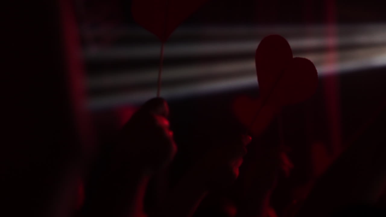 Hands holding red heart-shaped signs at a vibrant concert with dramatic lighting effects