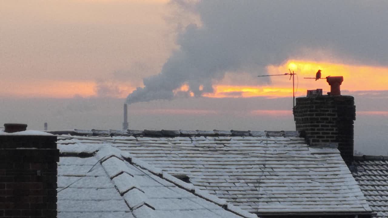 Smoking industry chimney above frosty winter home rooftops glowing sunrise orange sky