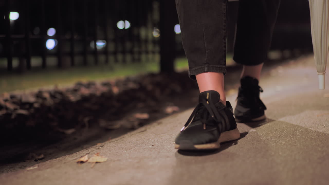 Close up of legs wearing black shoes and cropped pants holding umbrella while walking slowly along outdoor path covered with fallen leaves illuminated by soft street light during calm quiet night