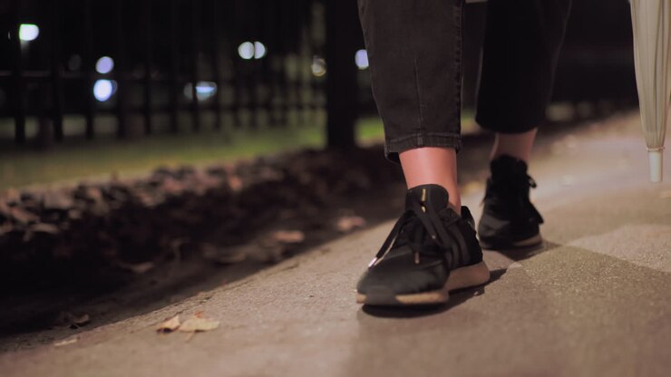 Close up of legs wearing black shoes and cropped pants holding umbrella while walking slowly along outdoor path covered with fallen leaves illuminated by soft street light during calm quiet night