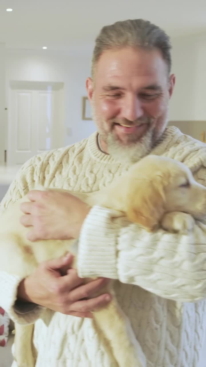 Video verticale: Uomo con un maglione che entra in cucina e culla un cucciolo di golden retriever, mentre una donna riprende.