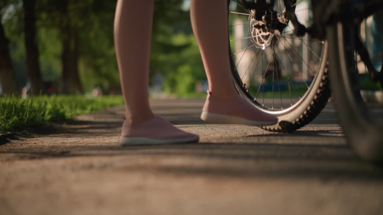 Close-up of individual legs in pink sneakers kicking a bicycle tire to check air pressure, lush greenery and trees create a beautiful backdrop with shadows on the ground with blurred background