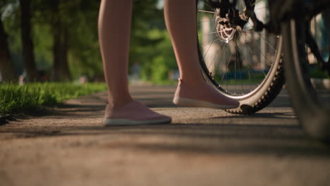 close-up of individual legs in pink sneakers kicking a bicycle tire to check air pressure, lush greenery and trees create a beautiful backdrop with shadows on the ground with blurred background