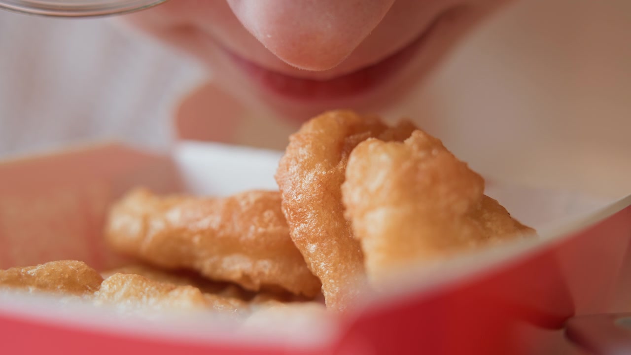 Close-up view of golden-brown battered fried chicken pieces in red paper takeout container while person leans in with hand and lower face nearby, suggesting hungry anticipation and imminent bite
