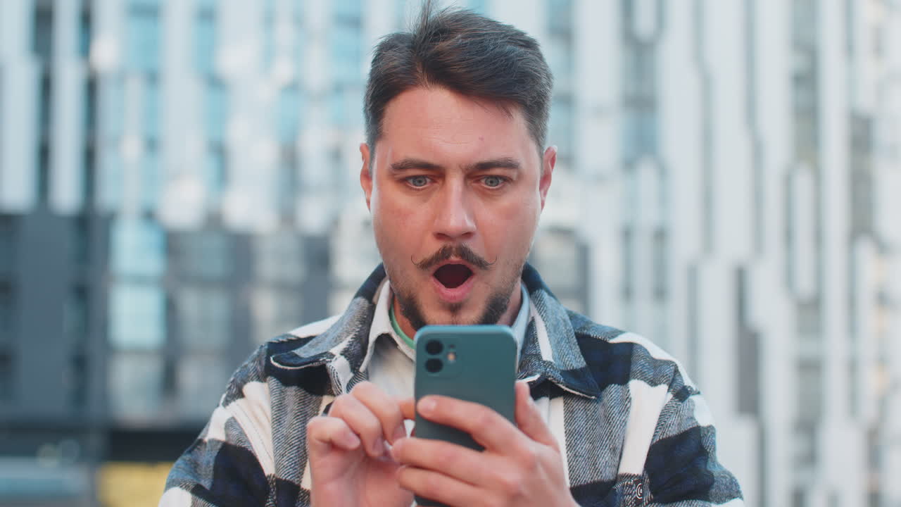 Caucasian mature man looking shocked while reading good news on smartphone in downtown city street