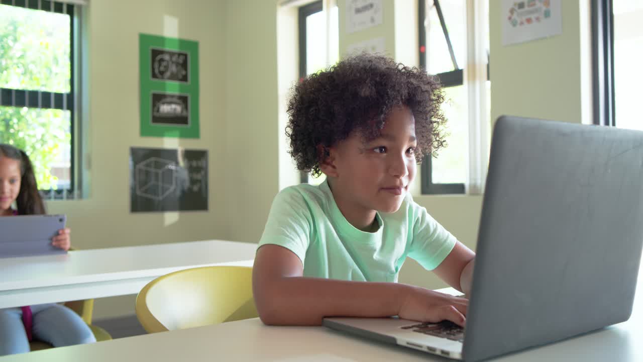 Ragazzo afroamericano che studia sul laptop al banco in classe, sorridendo mentre l'insegnante si avvicina per controllare il lavoro.