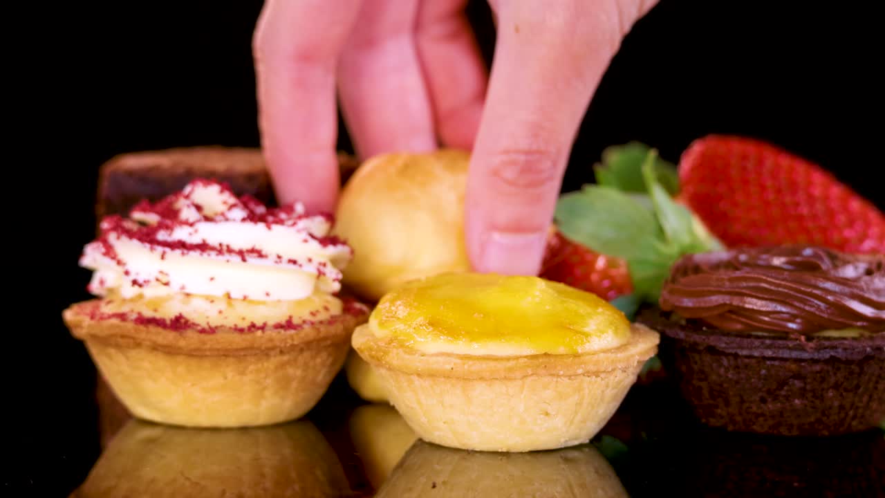 Hand selects a chocolate brownie from vibrant mini desserts on a reflective black surface