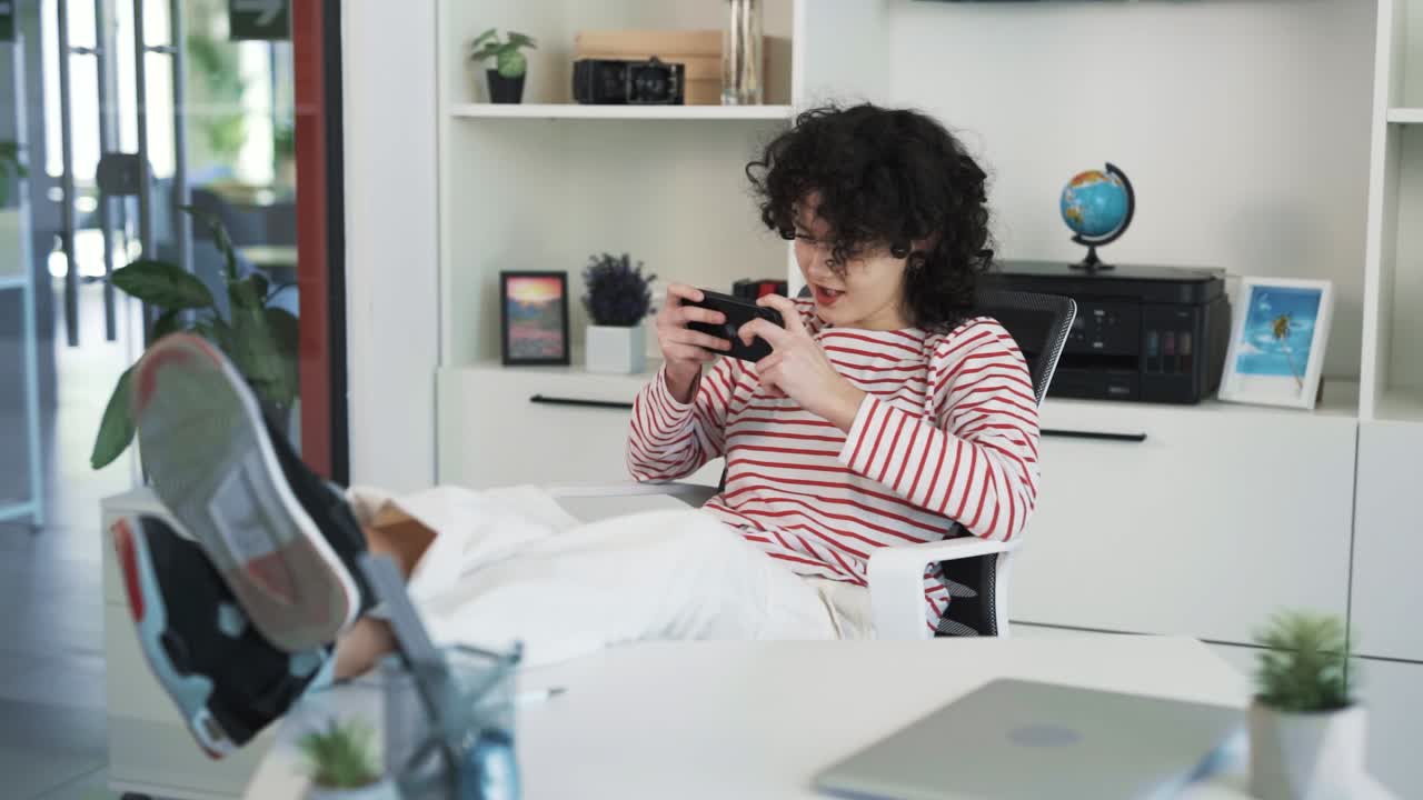 ritratto di una ragazza adulta dai capelli ricci emotivamente positiva seduta in un ufficio dietro una scrivania con i piedi sul tavolo, giocando a un gioco emozionante sul suo smartphone