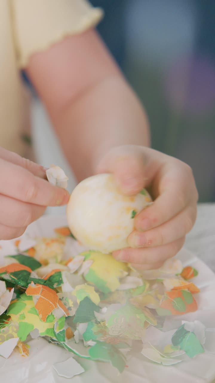 Child peels painted Easter egg gently. Hands focus on removing colorful shell pieces. Plate holds scattered confetti-like fragments. Scene feels calm and creative. Perfect for Easter, crafts