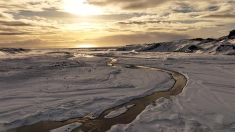 Vista-Aérea-Del-Paisaje-Sobre-Un-Río-Que-Fluye-Desde-El-Derretimiento-Del-Hielo-Del-Glaciar-Sólheimajökull,-El-Cambio-Climático,-En-Islandia,-Durante-La-Puesta-De-Sol