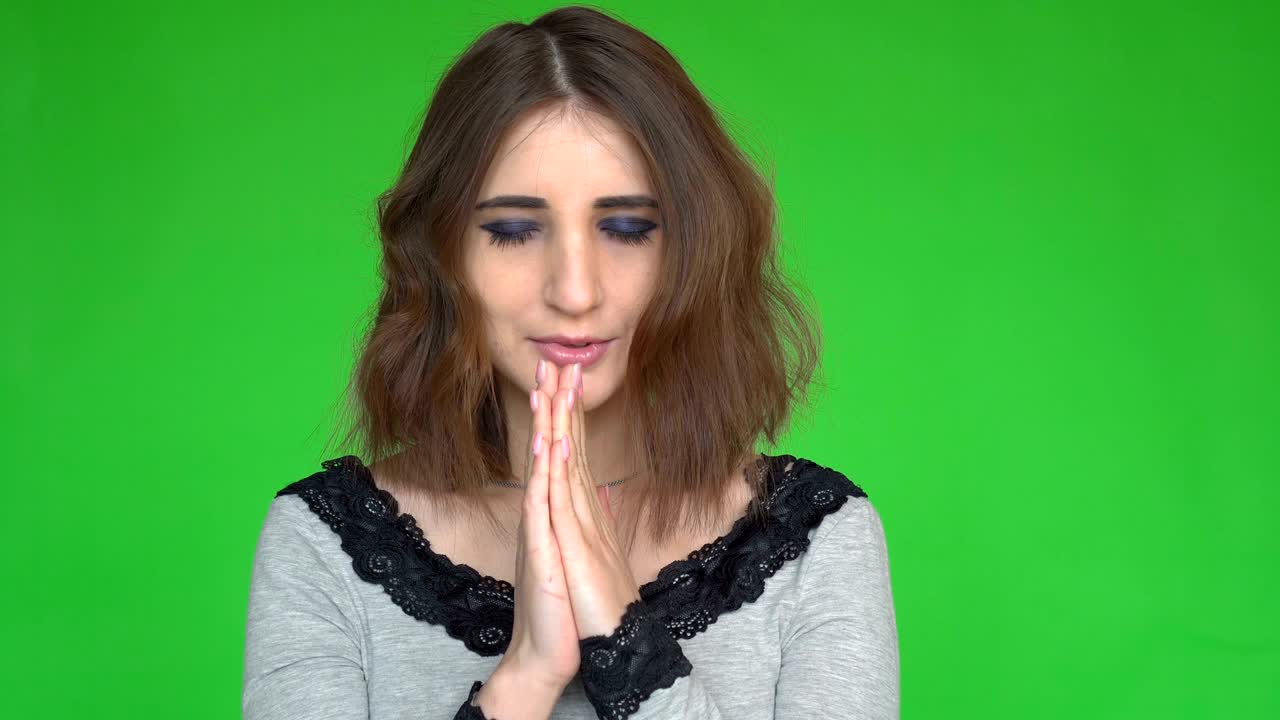 Hopeful female keeps hands together in praying gesture over green background.