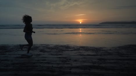 Slow-motion-landscape-view-of-a-silhouette-of-a-young-woman-running-on-a-sandy-beach-by-the-ocean,-at-sunrise