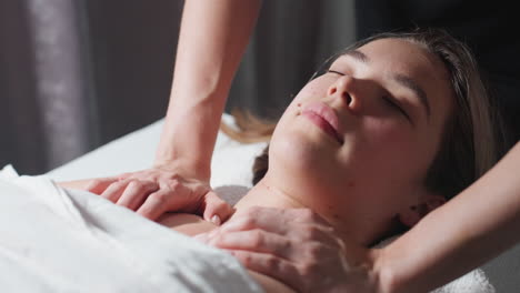 massage therapist in black uniform gently pressing collarbone area of light-skinned woman with closed eyes, lying peacefully on spa bed covered in white sheet under soft lighting