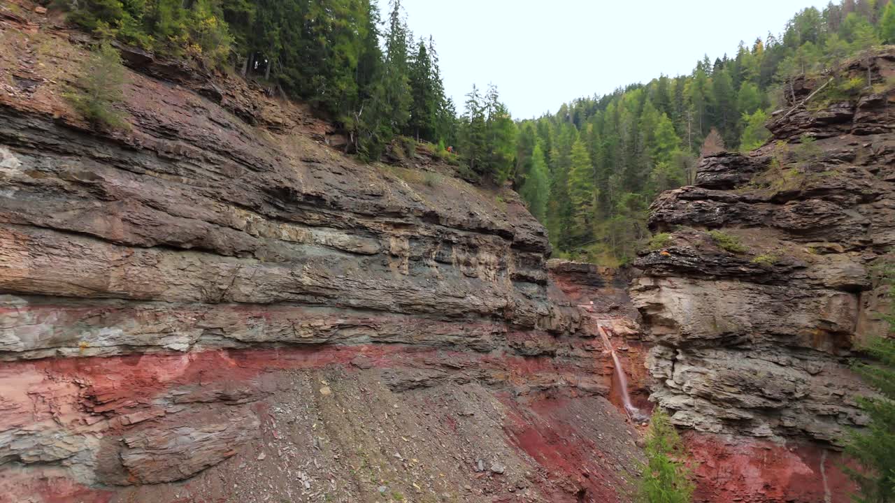 Slow motion reveal of a waterfall cascading down colorful, layered rock formations in Bletterbach Gorge, Italy. This rugged landscape highlights stunning geological features amidst lush forest.
