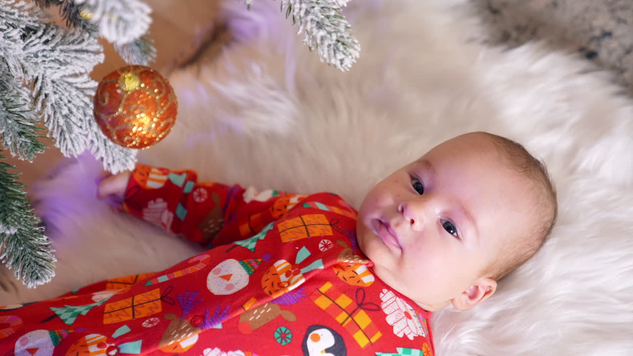 Cute toddler boy lying under the Christmas tree on a soft white plaid. Adorable child looks attentively up at a shiny decoration ball. Top view.