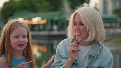 close up picnic scene mom with red manicure and daughter smiling as they playfully touch each other faces with paint brush near riverside at sunset with blurred greenery background