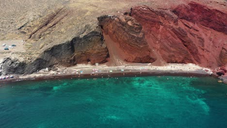 aerial drone flying towards red beach with turquoise water, boats, mountains and red colored sand in santorini, greece