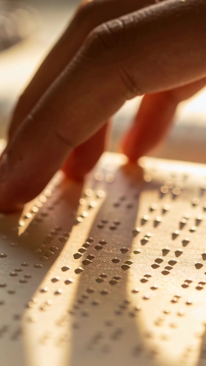 Vídeo vertical: Colocando uma única mão perto de página em braille na mesa, pontas dos dedos deslizando, lendo braille