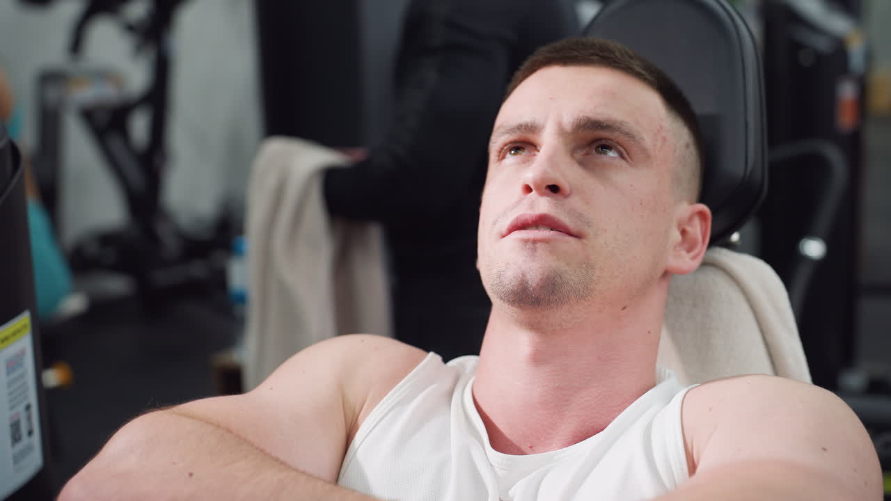 Student rests on leg press machine arms folded eyes lifted up reflecting recovery calm focus under modern gym environment with faint background patrons and equipment framing strength pause