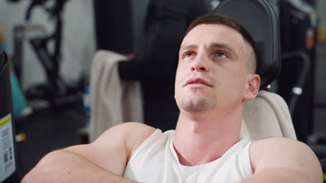 student rests on leg press machine arms folded eyes lifted up reflecting recovery calm focus under modern gym environment with faint background patrons and equipment framing strength pause